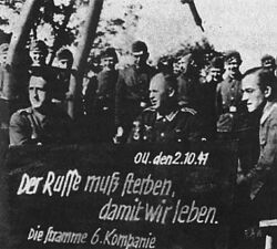 Вермахтісь военнослужащӧйезлӧн коллективнӧй фотография. Школаын велӧтанпӧв вылын мелӧн гижӧм: «Рочыслӧ колӧ кувны, мед олімӧ ми» (нем. Der Russe muß sterben, damit wir leben). Брянск область, 1941 во аршӧр 2 лун